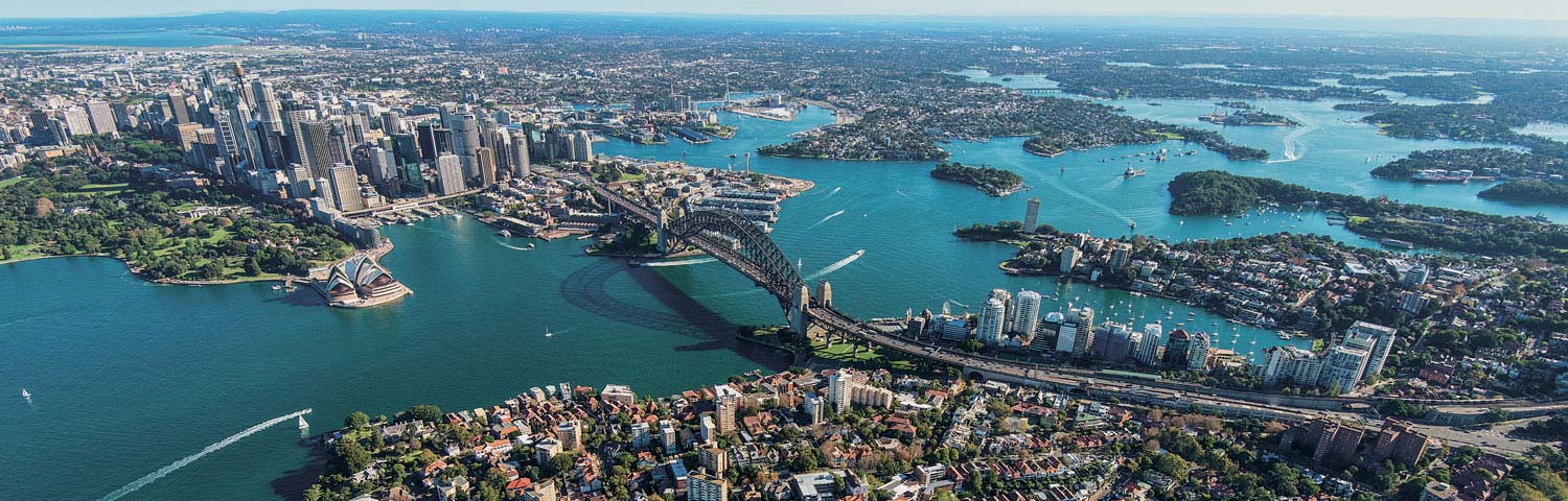 An aerial view of the Sydney Harbour Bridge over the water, and the surrounding city areas and ocean.